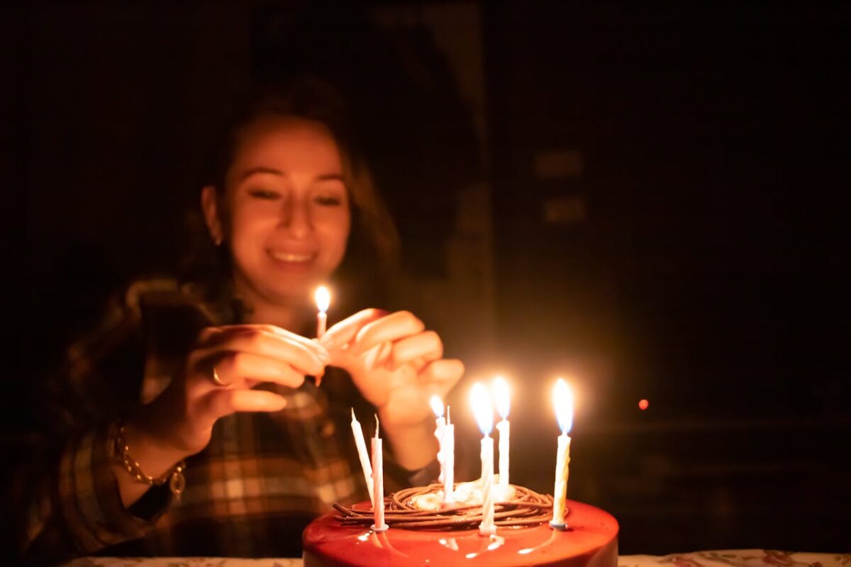 A joyful woman lighting candles on a birthday cake in a cozy, dimly lit environment.