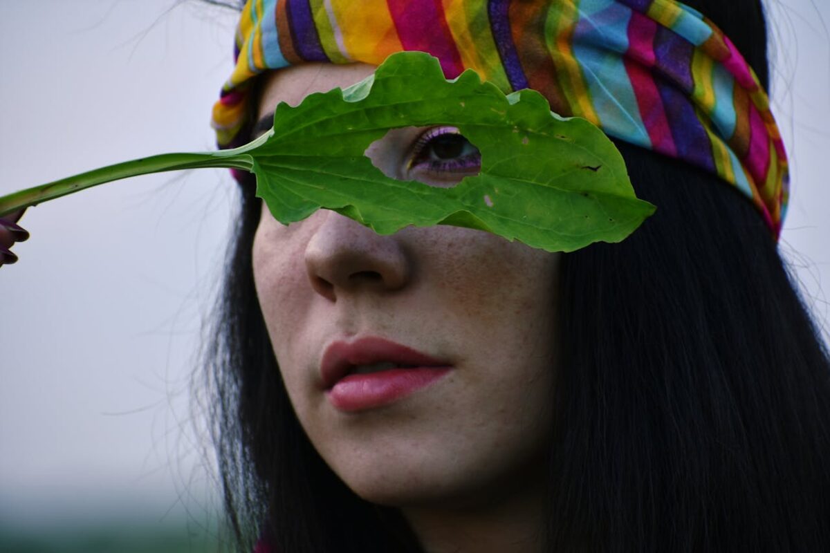 A woman with a colorful headband holds a leaf over her eye in a creative portrait.